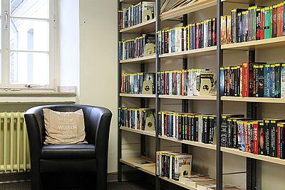 Ein schwarzer Sessel mit Kissen steht neben einem Wandregal voller Bücher und lädt zum verweilen ein